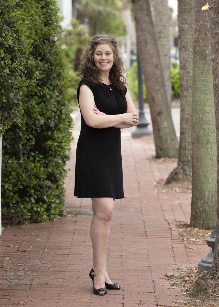 Suzanne, wearing black dress, standing outside on brick sidewalk with palm tree trunks and bushes in background.